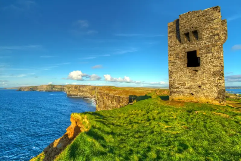 Medieval stone watchtower on dramatic Irish cliffs overlooking bright blue Atlantic Ocean with lush green countryside