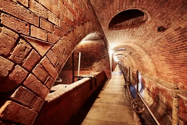 Historic underground sewer tunnel in Prague with red brick arched construction, featuring a narrow walkway with metal safety railings extending through the illuminated passage, showcasing the city's preserved medieval or early modern sanitation infrastructure.
