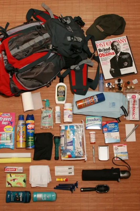 lat lay of travel and camping gear spread out on wooden floor including a red, black and gray hiking backpack, olive green military-style cap, Fast Company magazine, various toiletries and personal care items like sunscreen and hand sanitizer, travel-sized containers, medications, clear toiletry bags, small camping tools, and other outdoor adventure essentials organized for packing.