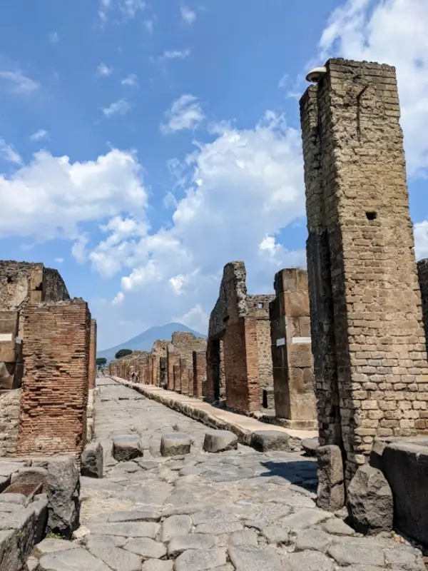 Ancient Roman street in Pompeii with weathered stone and brick walls lining both sides of a paved roadway, showing the preserved urban layout of the archaeological site with Mount Vesuvius visible in the distance under a blue sky with white clouds.