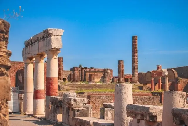 Ancient Roman ruins at Pompeii showing weathered stone columns, partial walls, and architectural remains scattered across the archaeological site, with Mount Vesuvius visible in the background under a clear blue sky.