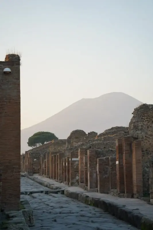 Ancient stone-paved street lined with Roman columns and ruined walls at the archaeological site of Pompeii, with Mount Vesuvius rising majestically in the background under a soft evening sky. The well-preserved ruins showcase the classic Roman architecture and urban planning of this ancient city, destroyed by the volcanic eruption in 79 AD.