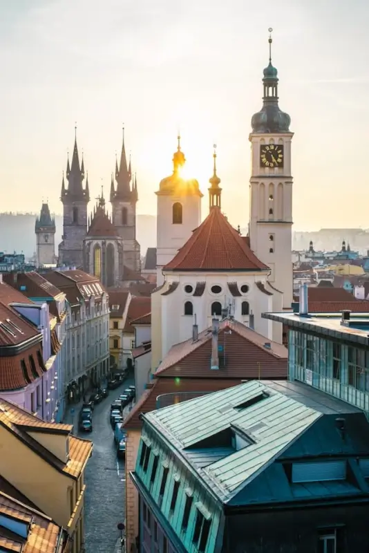 Panoramic view of Prague's historic Old Town at golden hour, showing the iconic bell tower with green dome and clock face in the foreground, Gothic church spires of Týn Church in the background, and traditional Czech architecture with red tile rooftops. The sun creates a warm glow behind the church domes, illuminating the medieval cityscape of Prague, Czech Republic.