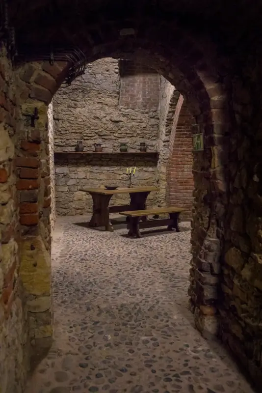 Historic underground chamber in Prague's tunnel system with arched brick and stone construction, featuring a wooden table with benches illuminated by candlelight, cobblestone flooring, and medieval masonry walls creating an atmospheric cellar space beneath the city.