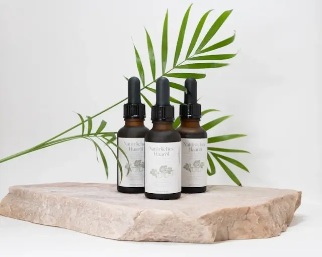 Three rosemary oil bottles with droppers displayed on natural stone platform with tropical palm leaves, ideal for travelers seeking portable wellness benefits