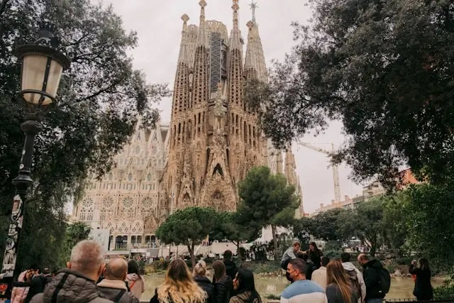 Ground-level view of Barcelona's Sagrada Família basilica showing the ornate Gothic and Art Nouveau facades with multiple towering spires featuring intricate stone carvings, framed by leafy trees on both sides, with a crowd of tourists gathered in the foreground and construction cranes visible indicating ongoing work on Gaudí's architectural masterpiece.
