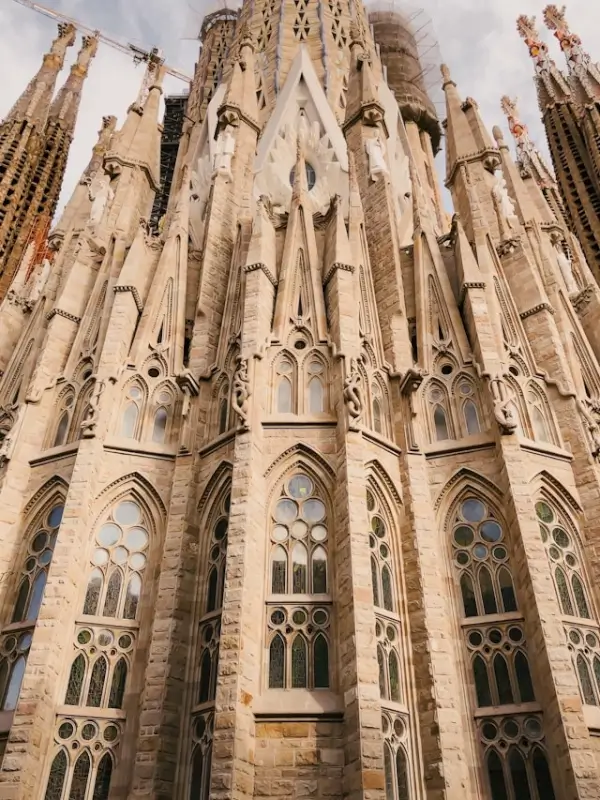 Low-angle view looking up at the Sagrada Família's intricately carved stone facade, showcasing multiple levels of Gothic arched windows with ornate tracery, towering spires with detailed sculptural elements, and elaborate stonework that demonstrates Gaudí's unique architectural vision, with construction cranes visible in the background against a cloudy sky.