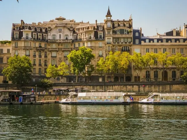 Seine River waterfront in Paris with traditional Haussmann buildings, green spring foliage, and tour boats moored along the stone embankment under clear blue sky
