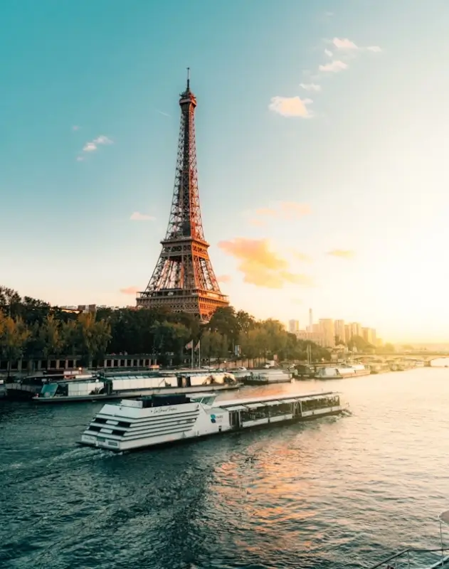 Eiffel Tower at golden hour with warm sunset light illuminating the iron lattice structure, viewed from the Seine River with tour boats and tree-lined embankment in foreground