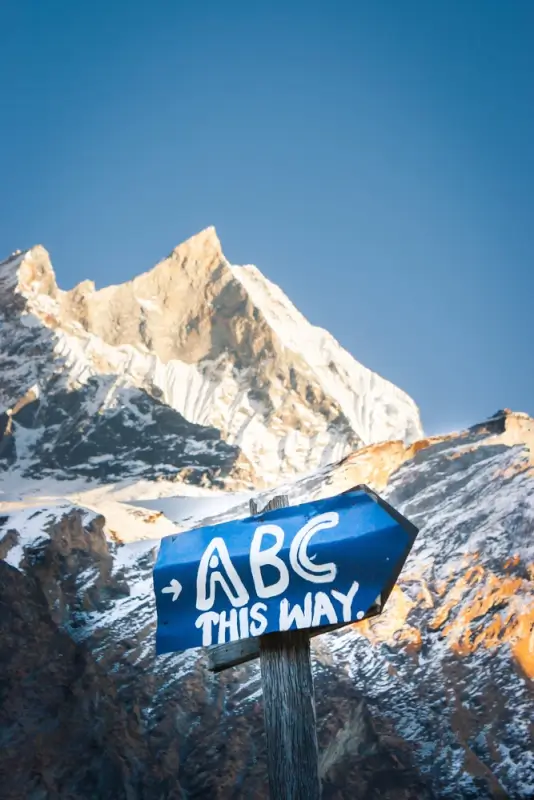 A blue directional sign reading 'ABC THIS WAY' points toward Annapurna Base Camp, with a towering snow-covered peak glowing in golden light against a clear blue sky in the background