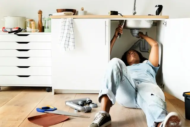 Person lying on kitchen floor working on plumbing repairs under the sink, with tools scattered nearby including wrenches and other equipment. The scene shows a modern kitchen with white cabinets, wooden countertop, and various kitchen items visible on the counter above.