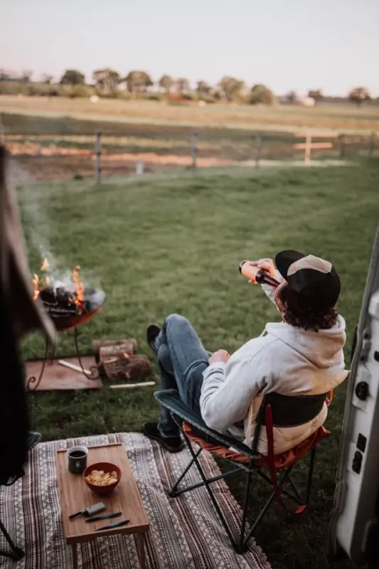 Person in hoodie and beanie relaxing in a camping chair beside their van, holding a mug while enjoying a campfire in a portable fire pit. The campsite is set up on grass with a patterned rug, camping supplies, and gear visible, overlooking a peaceful rural landscape with wooden fencing and rolling hills in the background during golden hour lighting.
