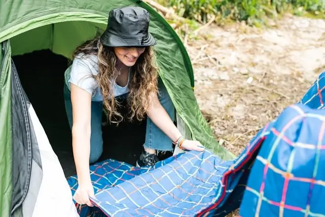 Woman with curly hair and dark hat smiling while setting up camping gear, arranging a blue plaid sleeping bag or blanket next to a green tent in an outdoor camping area with trees and natural ground cover visible in the background.