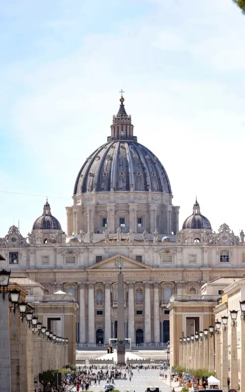 St. Peter's Basilica's massive dome and classical facade rise majestically above crowds of visitors gathered in St. Peter's Square, framed by the iconic curved colonnades
