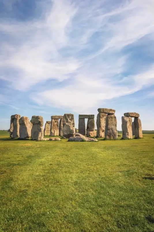 Stonehenge standing majestically on the green Salisbury Plain in England, showing the ancient stone circle with its iconic trilithons (two vertical stones topped by horizontal lintels) and standing sarsen stones arranged in their prehistoric formation, set against a dramatic sky filled with wispy white and blue clouds stretching across the expansive countryside landscape.