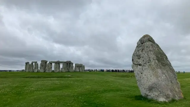 Wide-angle view of Stonehenge showing a large weathered sarsen stone in the foreground with the main prehistoric stone circle visible in the distance, surrounded by crowds of visitors on the vibrant green grass of Salisbury Plain, all set beneath a dramatic overcast sky with heavy gray and white clouds.