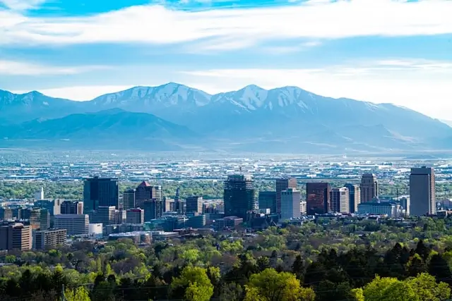 Panoramic view of Salt Lake City downtown skyline with the Wasatch Mountain Range in the background. The scene shows modern high-rise buildings and office towers in the city center, surrounded by green foliage and residential areas extending toward the dramatic snow-capped mountains under a partly cloudy blue sky.