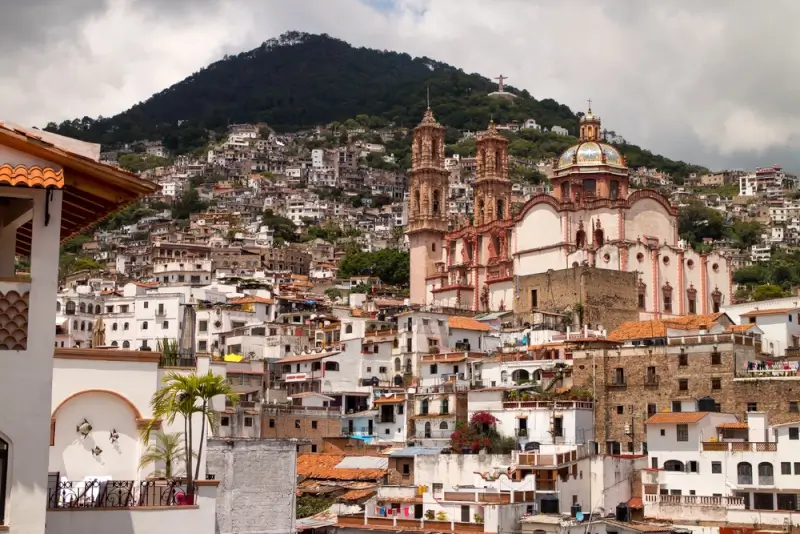 Historic Mexican colonial hillside town with baroque church featuring pink stone towers and dome, white buildings with terracotta roofs cascading up green mountainside under dramatic cloudy sky