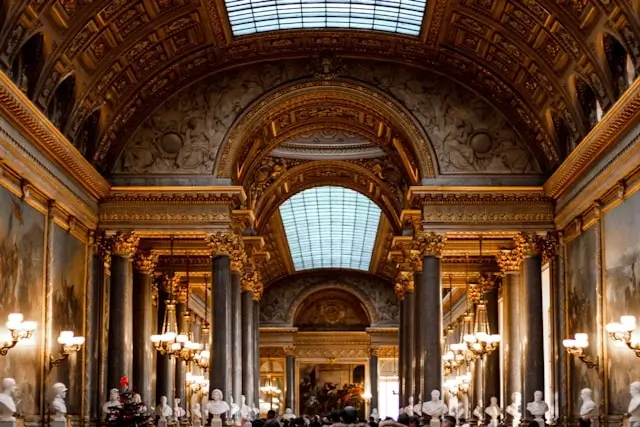 Ornate interior hall at Versailles featuring elaborate golden coffers on the vaulted ceiling, classical columns, crystal chandeliers, and glass skylights illuminating visitors below