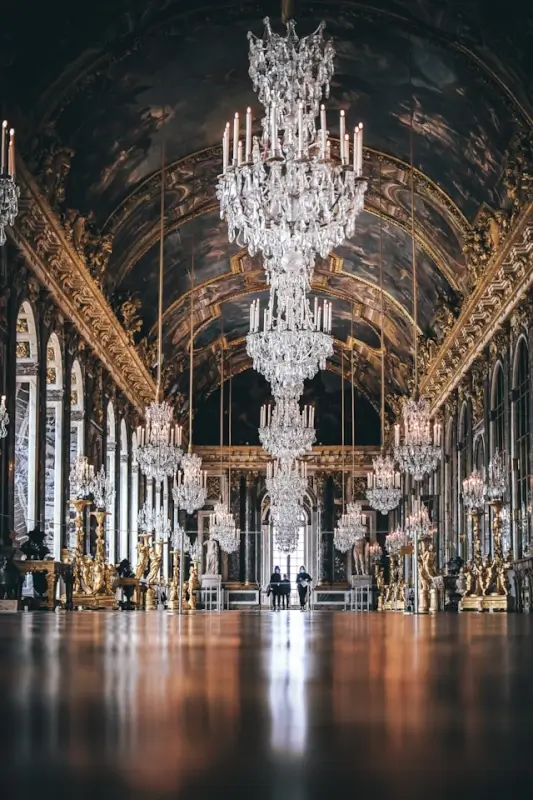 The magnificent Hall of Mirrors at Versailles with its famous gallery of crystal chandeliers suspended from an ornately painted vaulted ceiling, mirrored walls reflecting golden light, and polished parquet floors stretching the length of the grand hall
