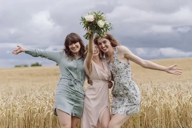 Three wedding guests in sage green, nude, and floral dresses posing joyfully in wheat field for outdoor ceremony