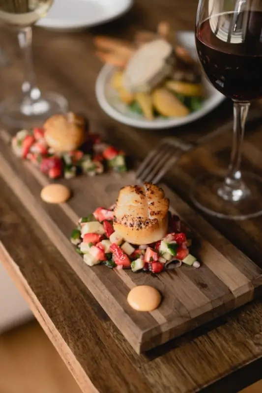 Gourmet seared scallops plated on a wooden board with colorful garnish, paired with a glass of red wine in an elegant restaurant setting