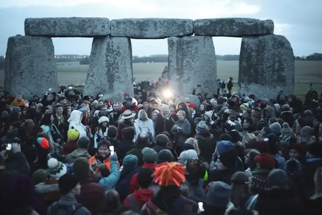 Large crowd of hundreds of people gathered at Stonehenge during what appears to be a solstice celebration, with visitors bundled in winter clothing densely packed around and within the ancient stone circle, while bright light (likely the sunrise or sunset) shines dramatically through one of the trilithon archways, creating a powerful alignment between the prehistoric monument and celestial event.