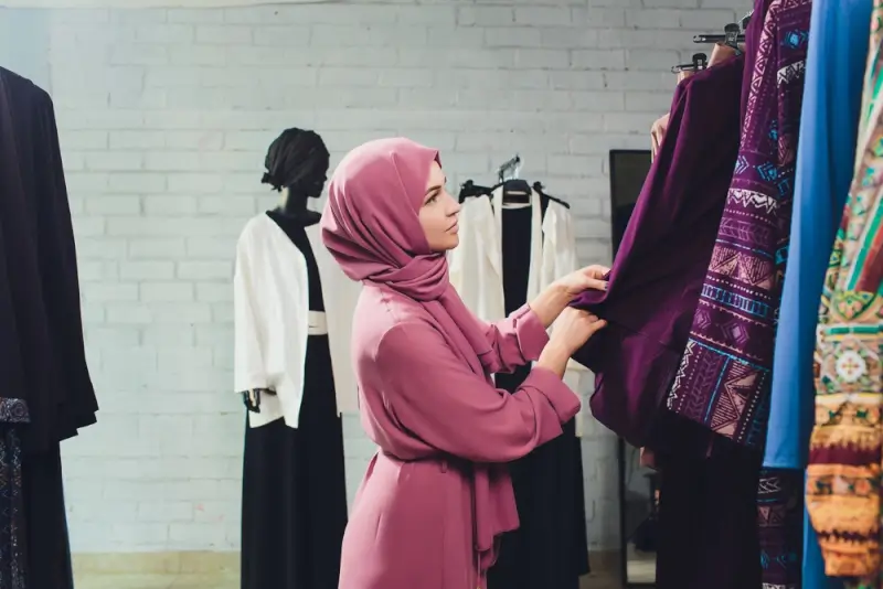 Hijabi woman choosing purple patterned abaya while wearing coordinated pink modest outfit
