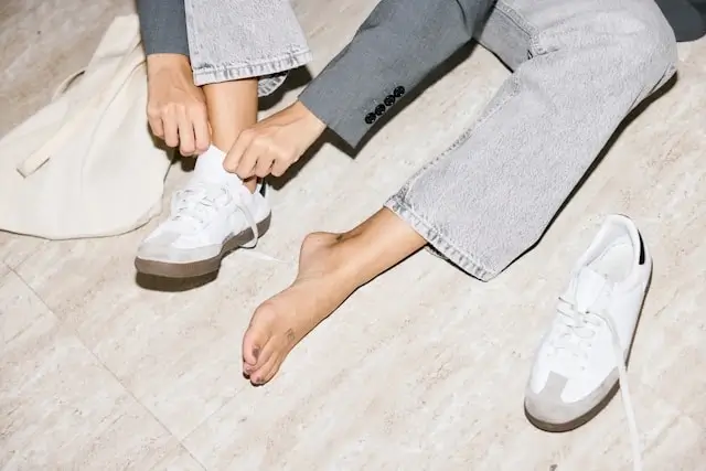 Person sitting and putting on white walking shoes while wearing light gray jeans on wooden floor