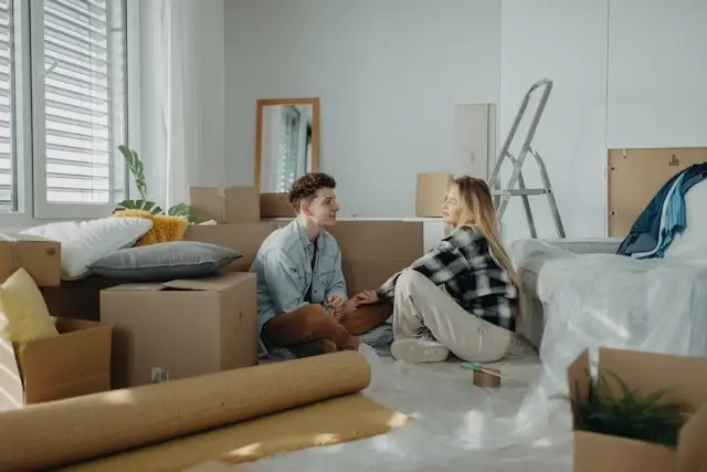 Young couple sitting on floor surrounded by moving boxes and belongings in minimalist apartment during relocation process
