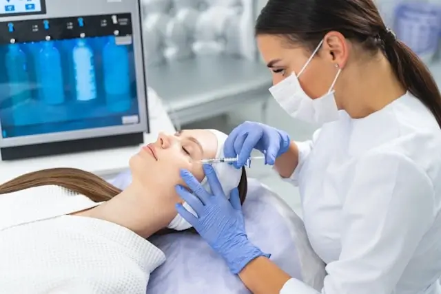 Medical professional in white coat and face mask administering an injection to a patient's face using a syringe, with medical equipment and monitor visible in the background.
