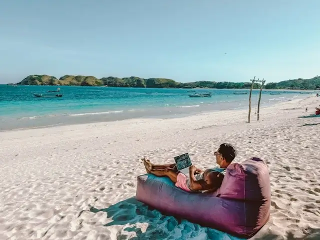 Digital nomad working on laptop from tropical beach with turquoise water, demonstrating remote work and adventure travel lifestyle
