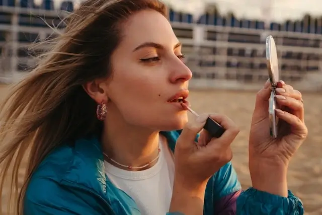 Woman applying lipstick while looking in handheld mirror outdoors, wearing blue jacket with wind-blown hair in golden hour lighting