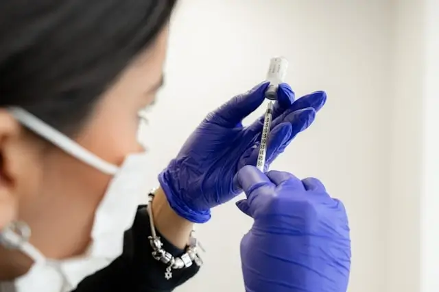 A professional dermatologist wearing a face mask and blue medical gloves preparing a syringe, viewed from below against a white ceiling.