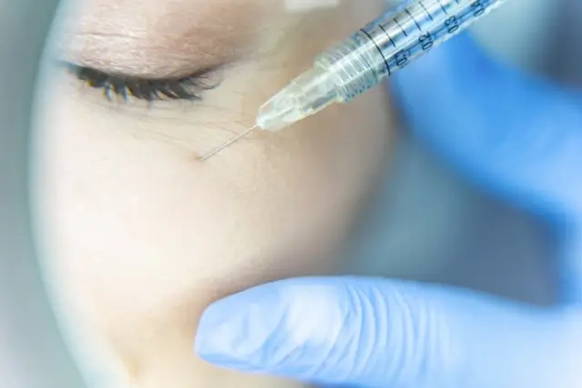 Extreme close-up of a syringe needle positioned near the eye area during a precise cosmetic injection procedure, with blue gloved hand visible.