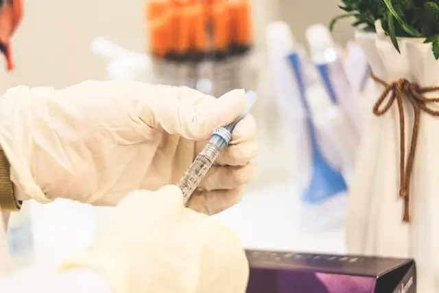 Close-up of a gloved hand holding a medical syringe in a clinical setting with medical supplies and a small plant visible in the background.