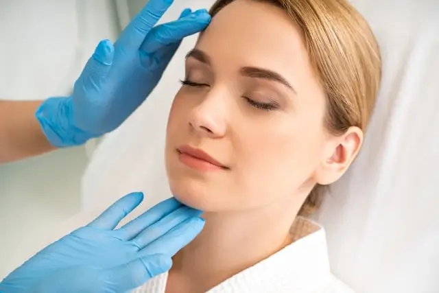 Woman with closed eyes lying down while practitioner's hands in blue medical gloves examine her face and forehead area during a consultation.