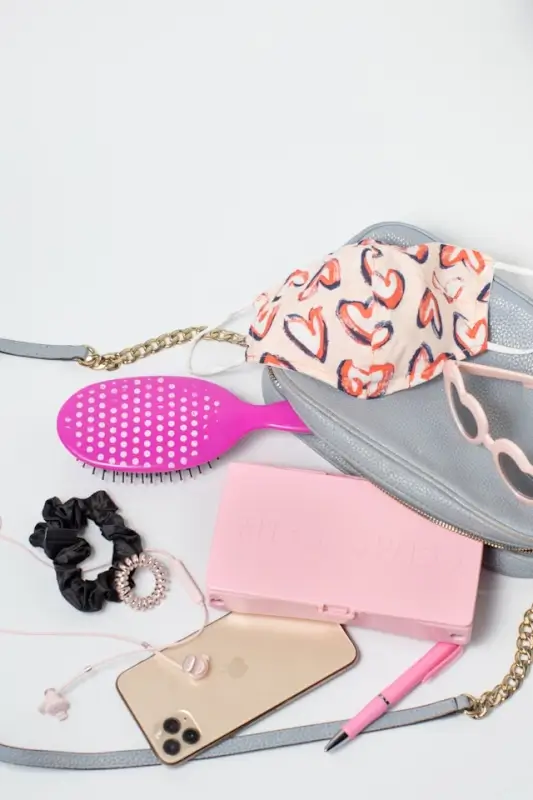 Flat lay of women's accessories including gray crossbody bag, heart-patterned face mask, pink detangling brush, black hair scrunchies, pink wallet, beige phone case, and pink pen on white background