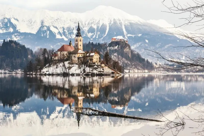 Lake Bled church on island with snow-capped Julian Alps reflected in calm winter waters, showcasing Slovenia's iconic outdoor destination and gateway to Balkan adventures