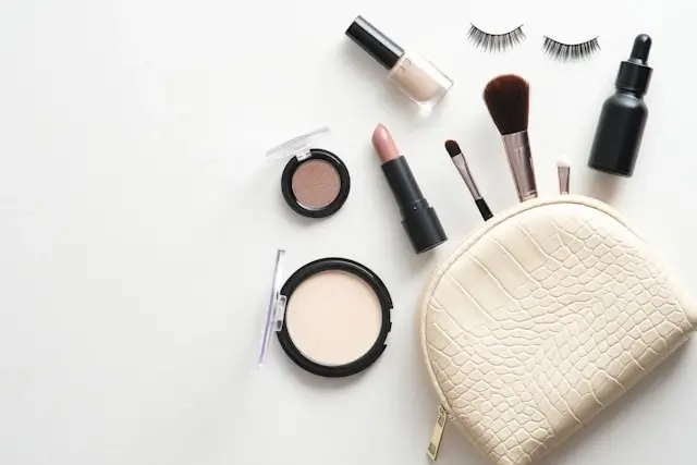 Flat lay of makeup products including lipstick, eyeshadow, foundation, makeup brushes, false eyelashes, and serum arranged around cream quilted cosmetic bag on white background