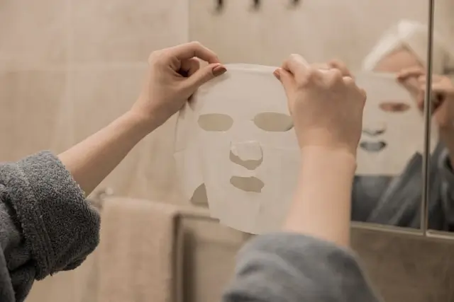 Woman applying white sheet face mask in front of bathroom mirror, hands positioning mask on face