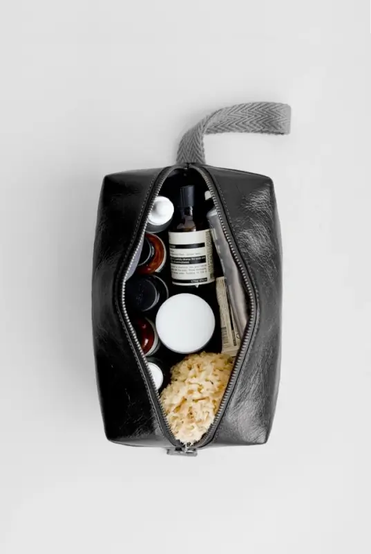 Open black leather toiletry bag with gray herringbone strap containing various skincare products including bottles with pump dispensers, jars with white lids, and what appears to be a natural loofah or sponge, arranged on a light gray background.