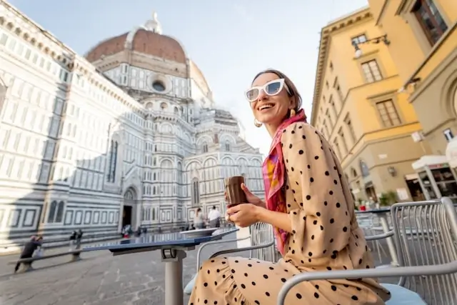 Smiling woman wearing sunglasses and a beige polka dot dress with red scarf sits at an outdoor café table in front of Florence Cathedral (Duomo) with its distinctive Renaissance dome and ornate marble facade visible in the background.