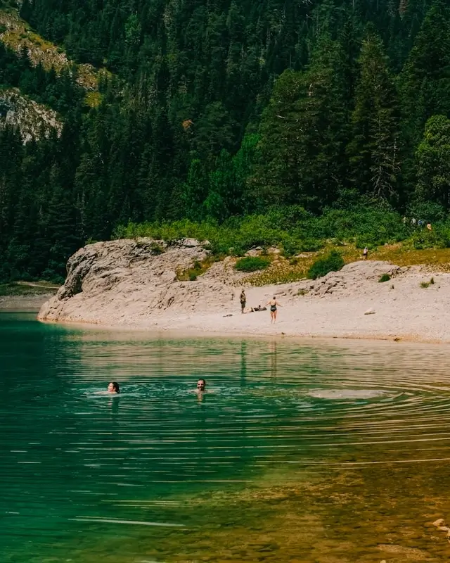 Swimmers enjoying crystal-clear turquoise lake waters surrounded by dense forest in the Balkans, with sandy beach and dramatic cliffs creating perfect outdoor adventure destination