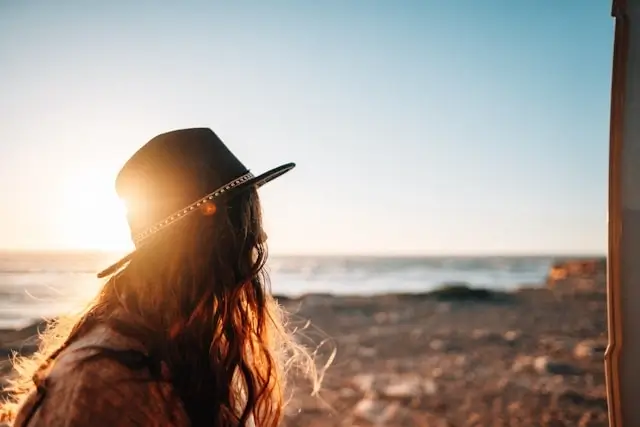Woman with long wavy hair wearing a dark hat with decorative band, silhouetted against golden sunset light on a beach with ocean waves and sandy shore in the background.