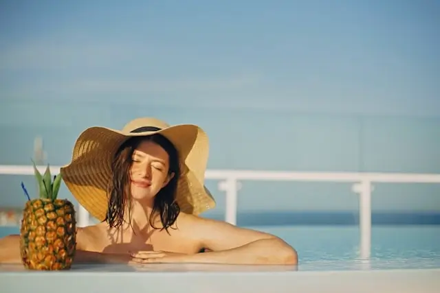 Woman relaxing in infinity pool wearing wide-brimmed straw hat with tropical pineapple drink, ocean view in background