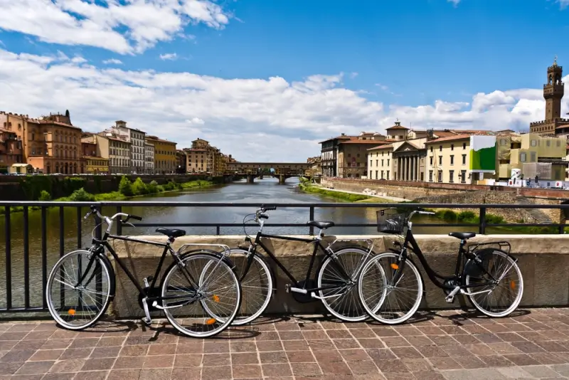 Cycling in Italy through Florence with bikes parked along the Arno River overlooking historic Ponte Vecchio and Renaissance architecture