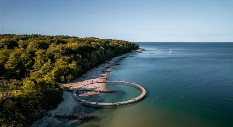 Aerial view of the Infinite Bridge in Aarhus Bay, Denmark, showing the circular walkway extending into the Baltic Sea with forested coastline and beach