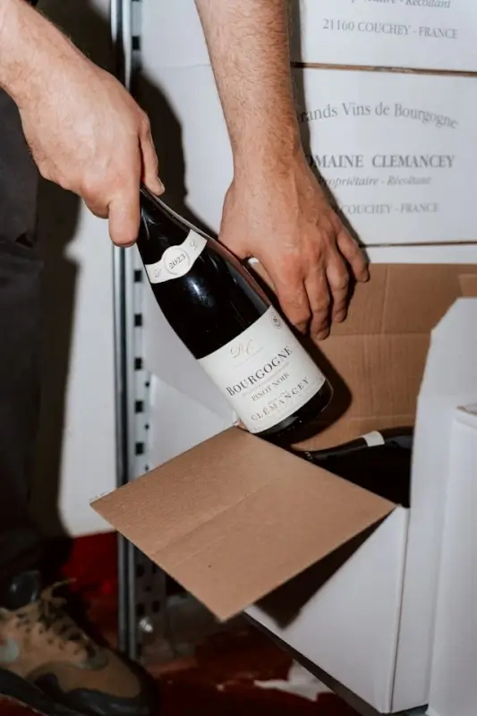 Hands carefully placing a 2020 Bourgogne Pinot Noir wine bottle from Domaine Clemancey into a cardboard shipping box, with wine storage boxes visible in background