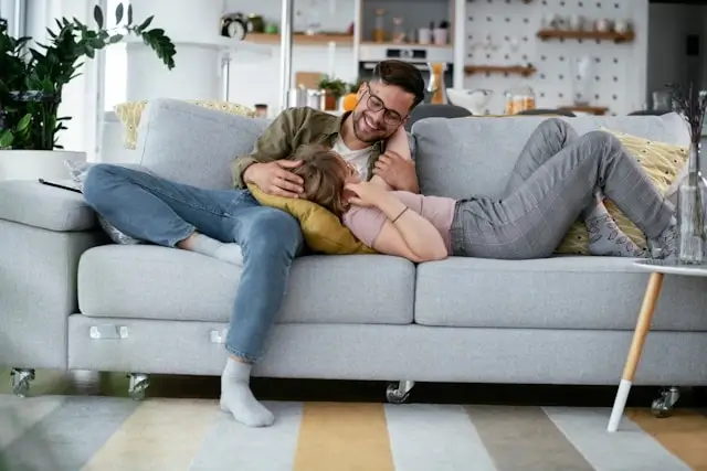 Happy couple cuddling intimately on gray couch in modern living room, showing emotional connection and bedroom confidence between partners